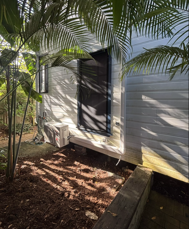 Outdoor unit installed beneath a raised weatherboard home, shaded by palms