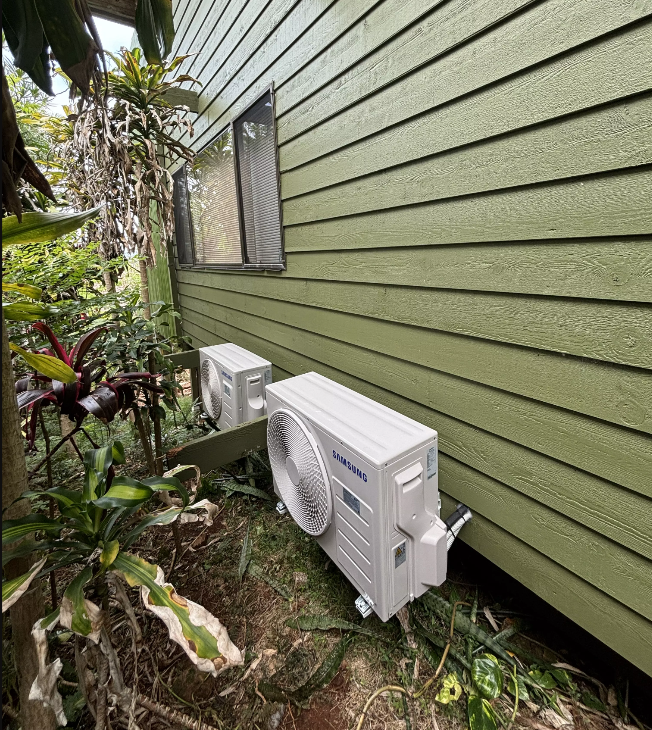 Two Samsung outdoor units installed against a green weatherboard wall among garden plants